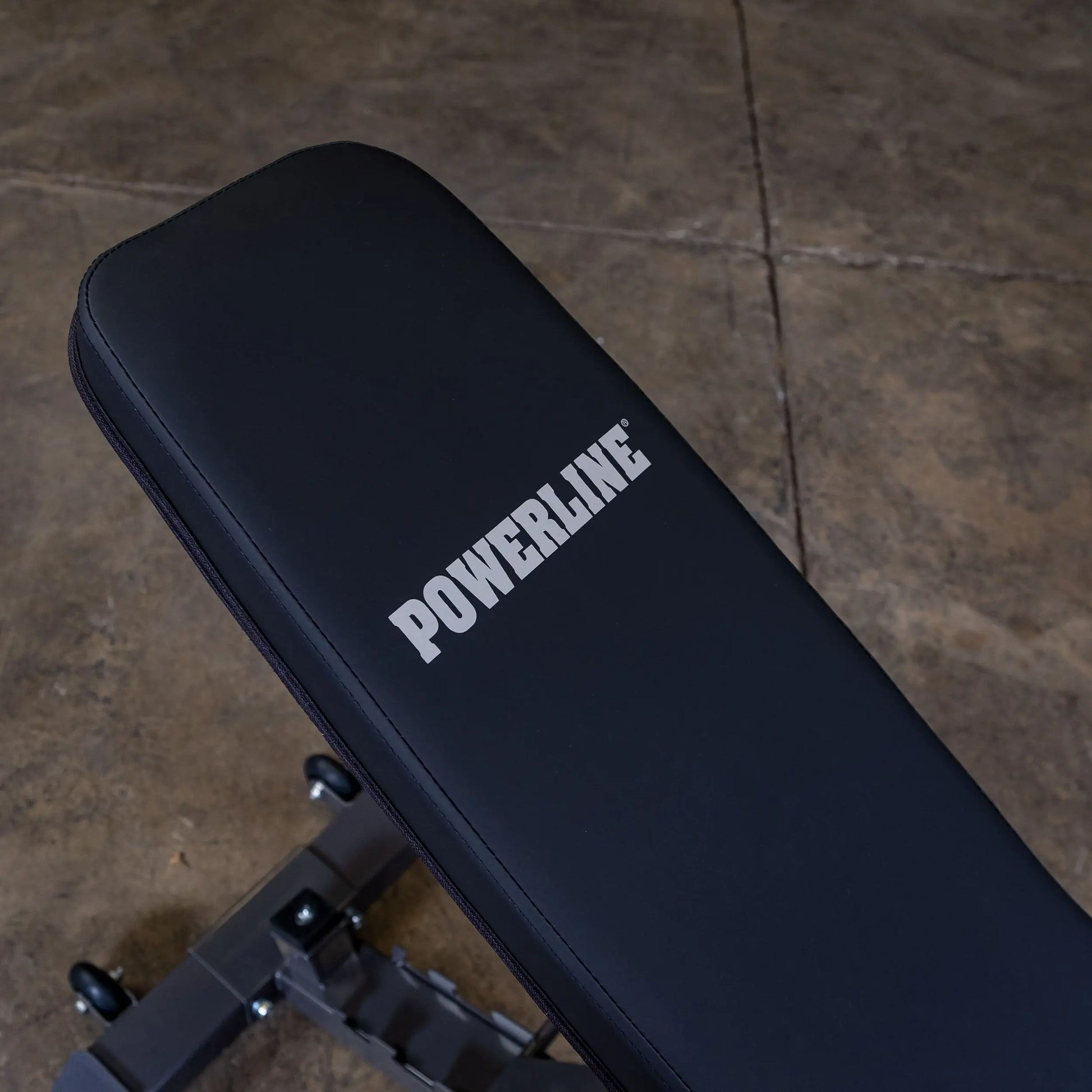 Close-up of a black workout bench with the word POWERLINE prominently displayed in white on the padded surface. The bench is positioned on a concrete floor.