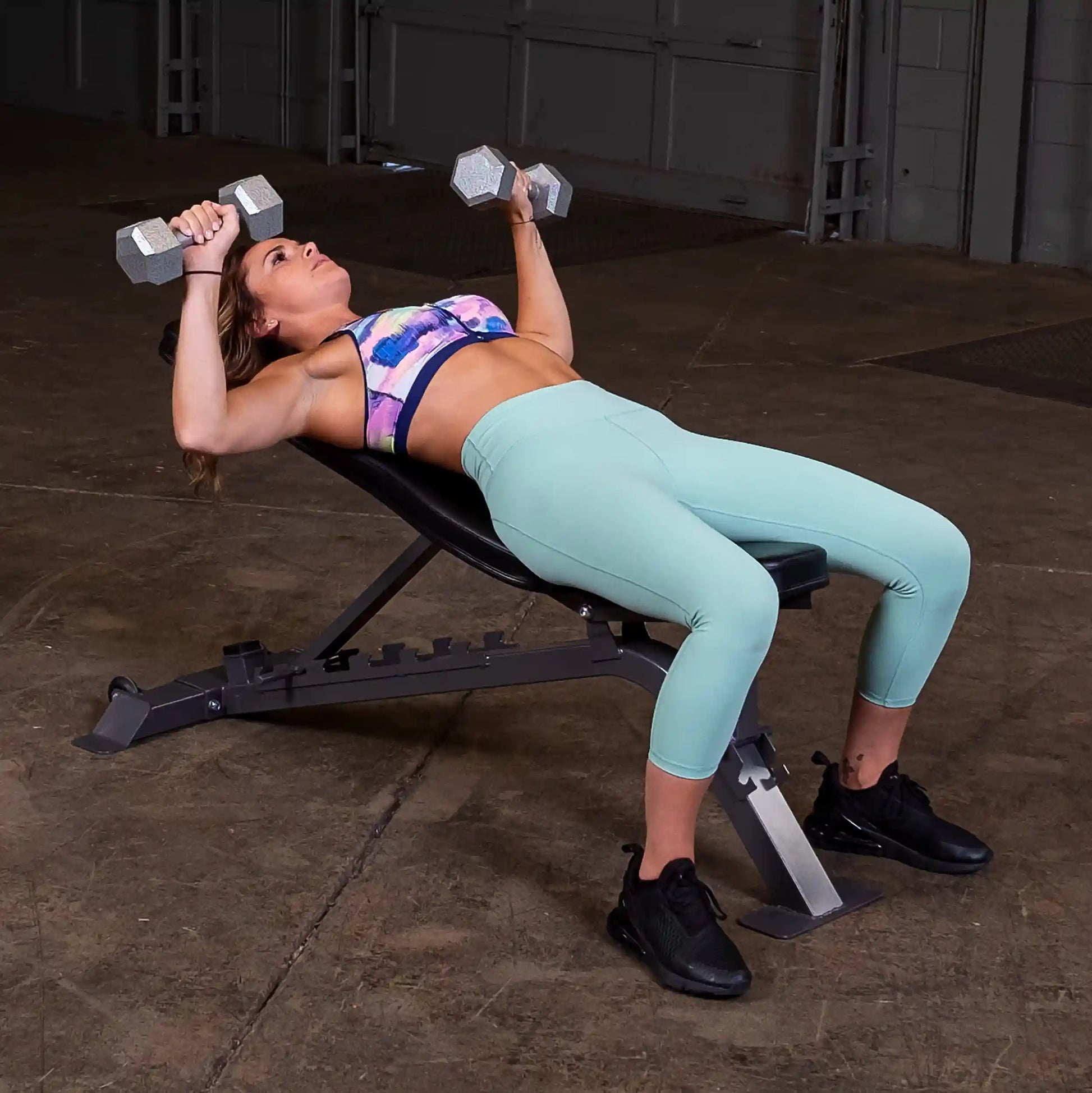 A woman lying on a workout bench, performing a dumbbell press with weights in both hands. She is wearing a colorful sports bra and light blue leggings, with black athletic shoes. The background shows a gym environment with concrete flooring.