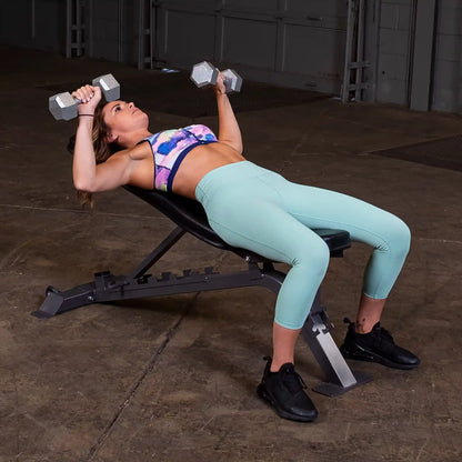 A woman lying on a workout bench, performing a dumbbell press with weights in both hands. She is wearing a colorful sports bra and light blue leggings, with black athletic shoes. The background shows a gym environment with concrete flooring.
