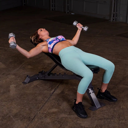 A woman performing a chest exercise on a weight bench, lying back with dumbbells in both hands, wearing a colorful sports bra and light blue leggings in a gym setting.