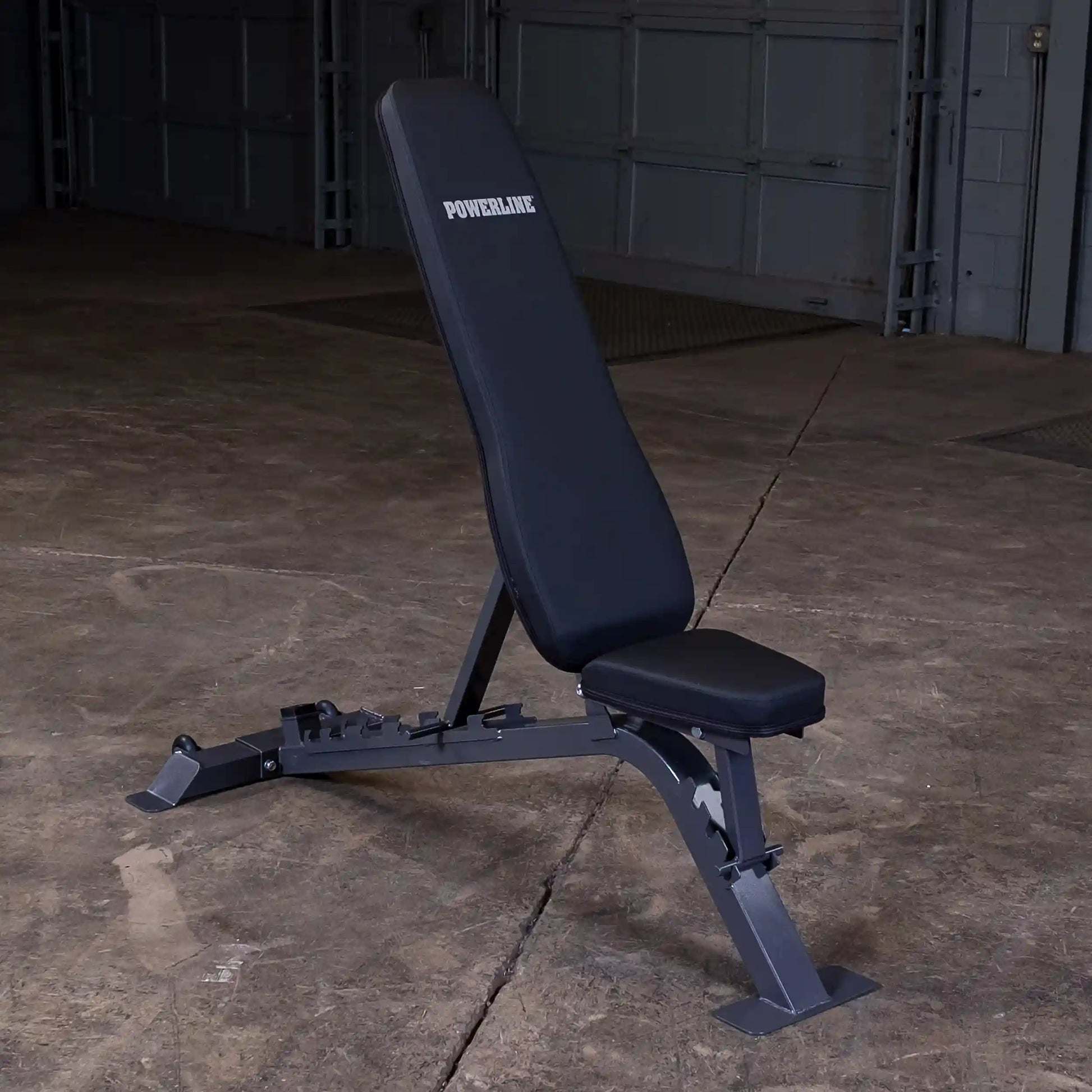 A black adjustable weight bench with the brand name POWERLINE displayed on the backrest, set against a concrete floor in a dimly lit garage.