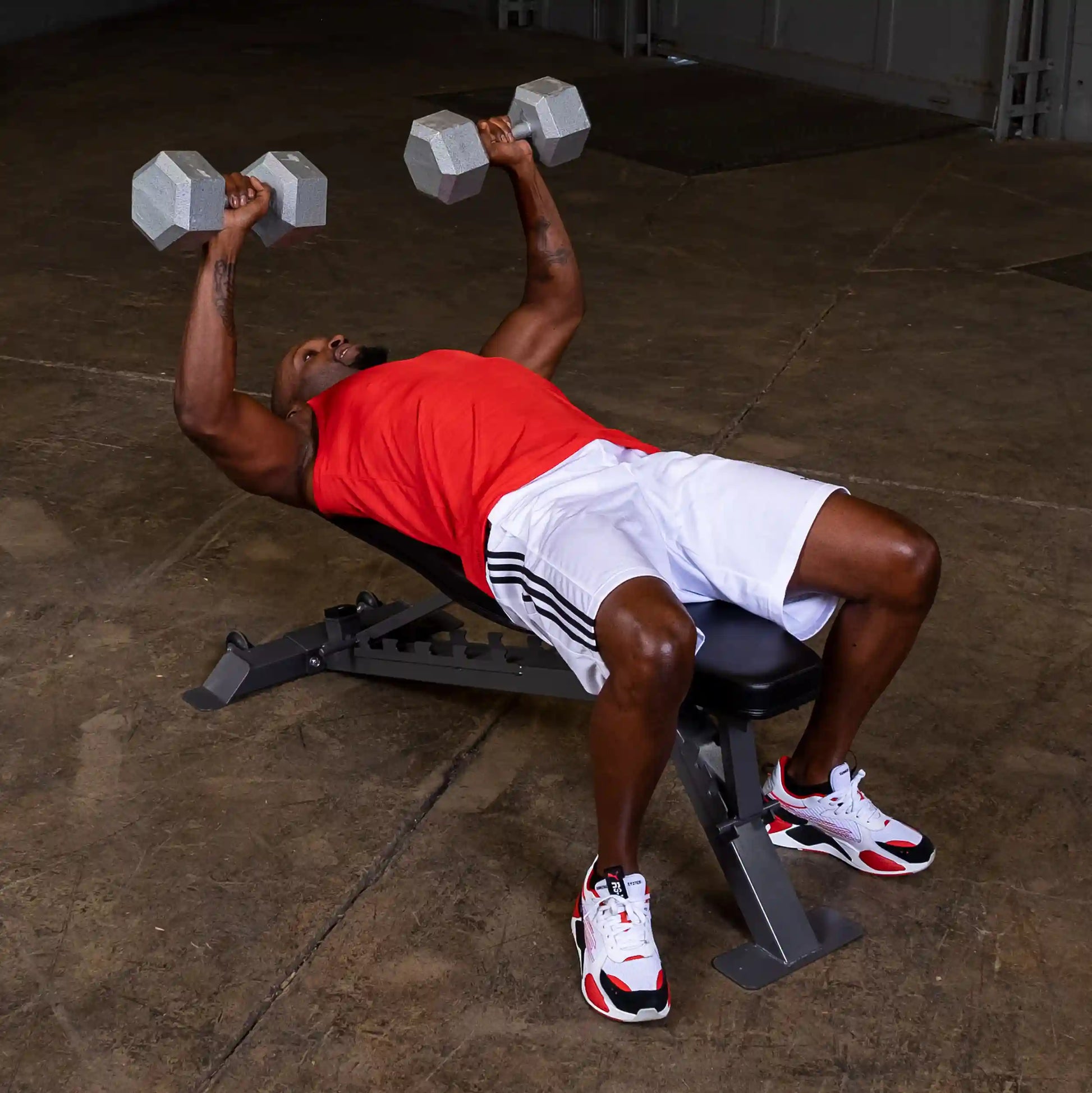 A man in a red tank top and white shorts performs a dumbbell bench press on a workout bench in a gym setting. He is lifting two gray dumbbells above his chest with a focused expression.