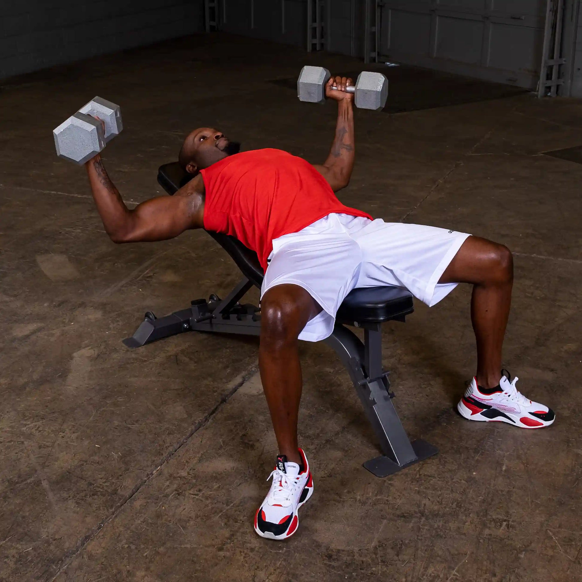 A person performing a dumbbell chest press on a bench, wearing a red sleeveless shirt and white shorts, with weights in both hands in a gym setting.