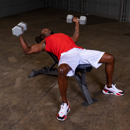A person performing a dumbbell chest press on a bench, wearing a red sleeveless shirt and white shorts, with weights in both hands in a gym setting.