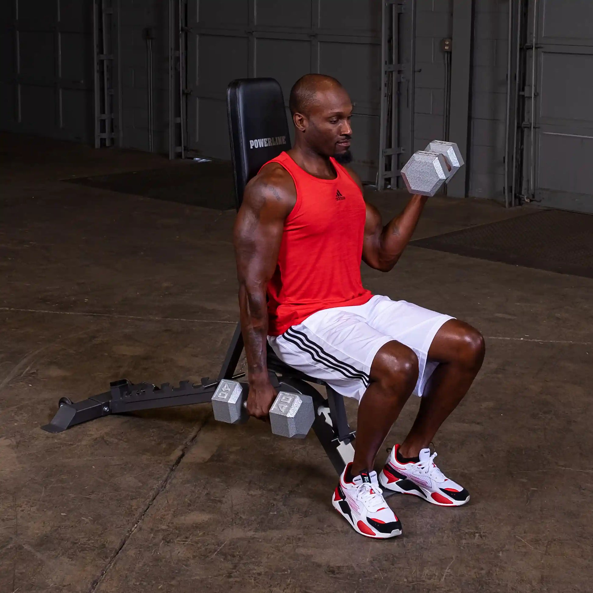 A muscular man in a red tank top and white shorts seated on a weight bench, performing a dumbbell bicep curl with a pair of gray dumbbells in a gym setting.