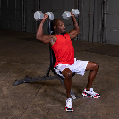 A muscular man in a red tank top and white shorts is seated on a weight bench, lifting dumbbells overhead in a gym setting.