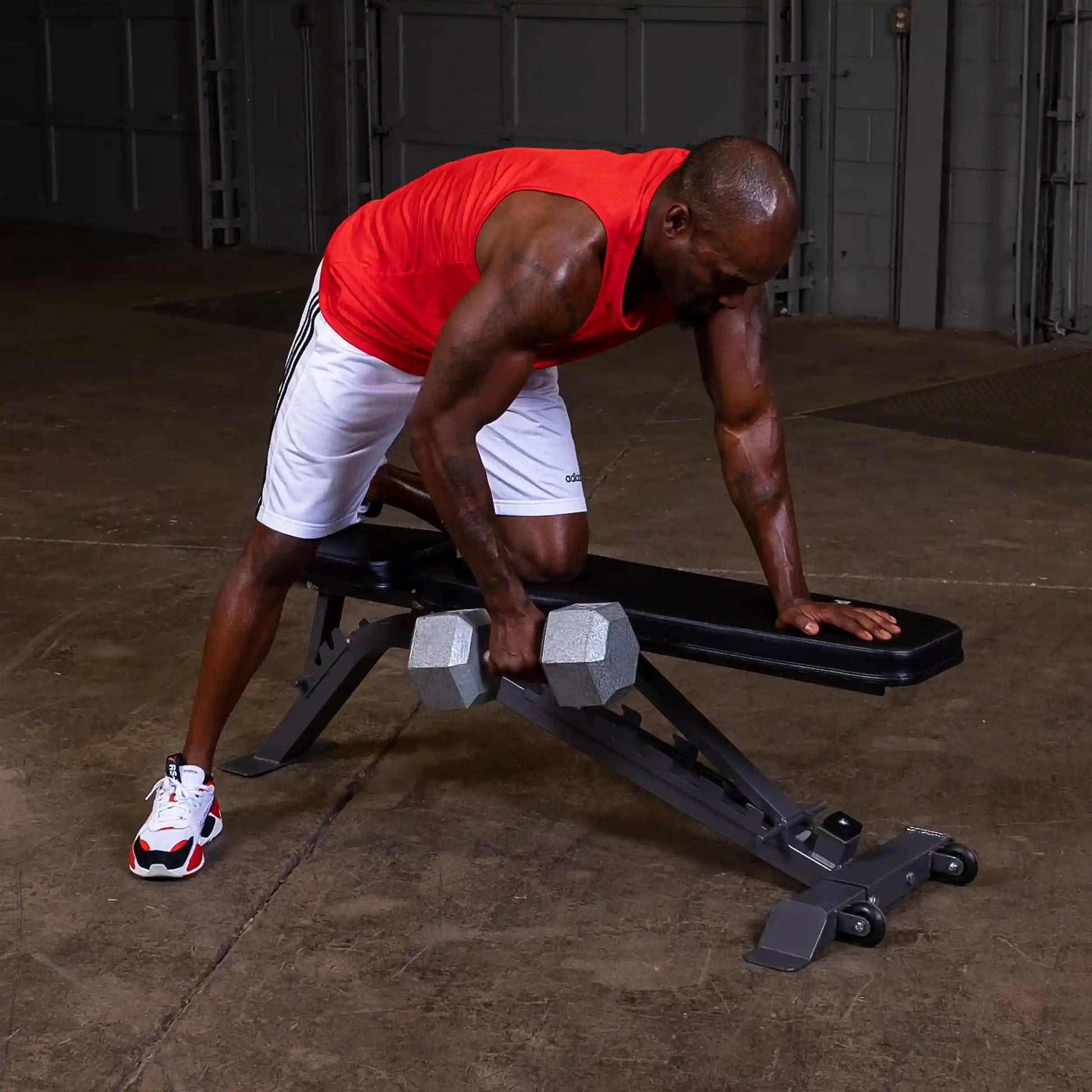 A person in a red tank top and white shorts performs a single-arm row exercise with a dumbbell on a workout bench in a gym setting.