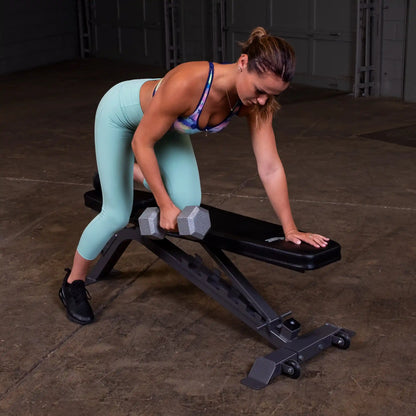 A woman in a colorful sports bra and mint green leggings performs a bent-over dumbbell row on a weight bench in a gym setting.