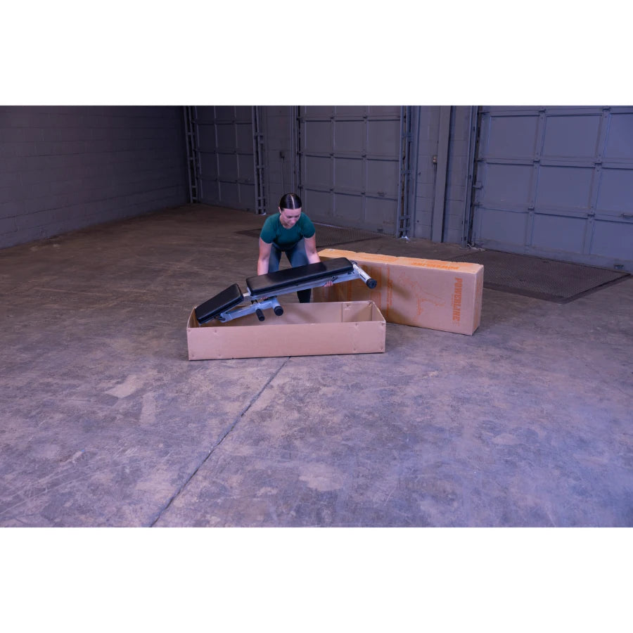 A person unpacking a weight bench from a cardboard box in a spacious indoor area with a concrete floor and garage doors in the background.
