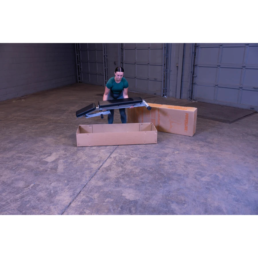 A woman in a green shirt is lifting a piece of equipment out of a cardboard box in a large, empty space with concrete flooring.