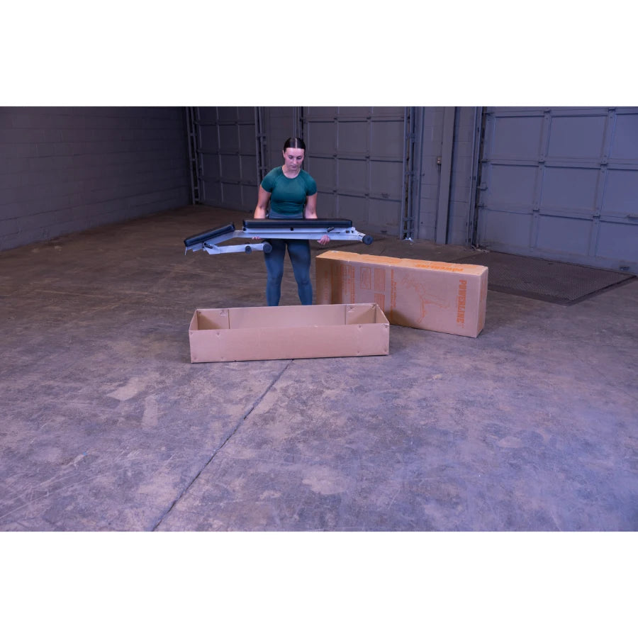A woman in a green shirt is holding a folding table above a cardboard box, with another box visible in the background, inside a spacious garage.