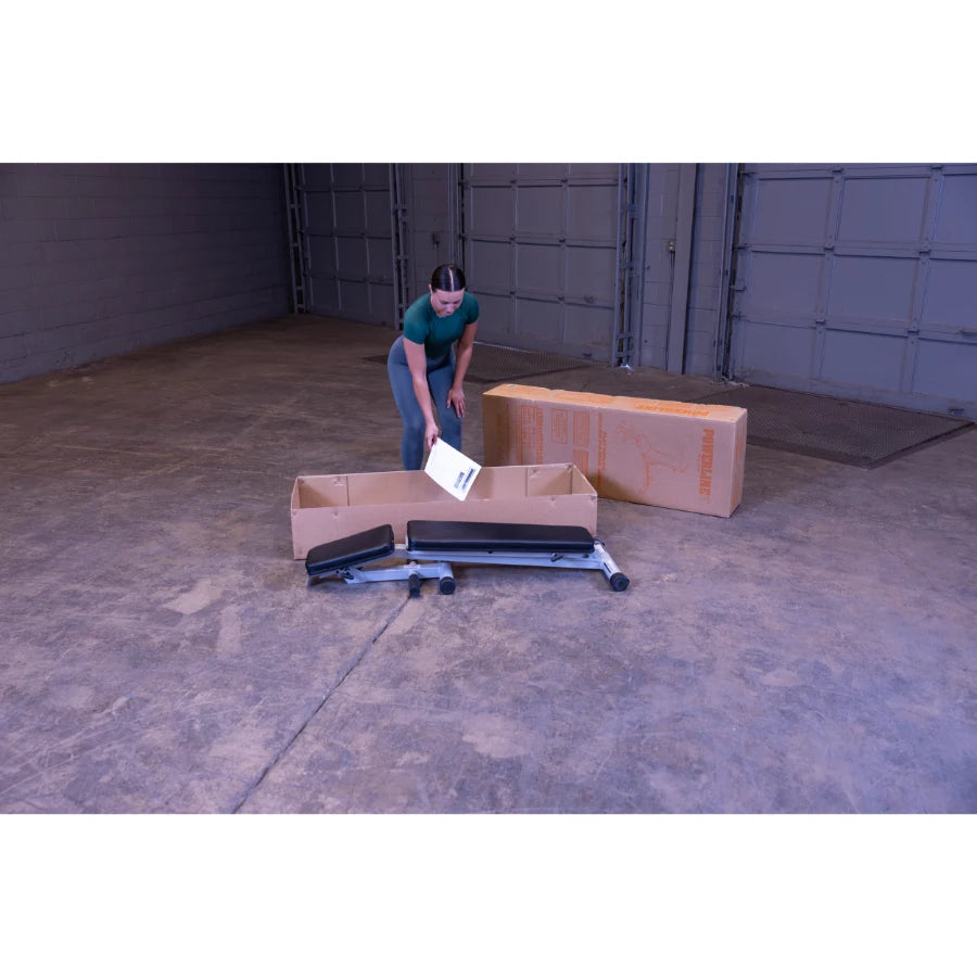 A person in a green shirt and gray leggings is unpacking items from a large cardboard box in a spacious, empty room, with a partially assembled piece of equipment and a manual on the floor.