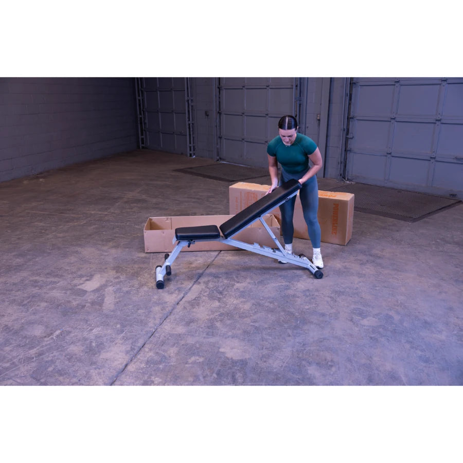 A person in athletic wear adjusting a weight bench in a spacious, industrial-style room with cardboard boxes in the background.