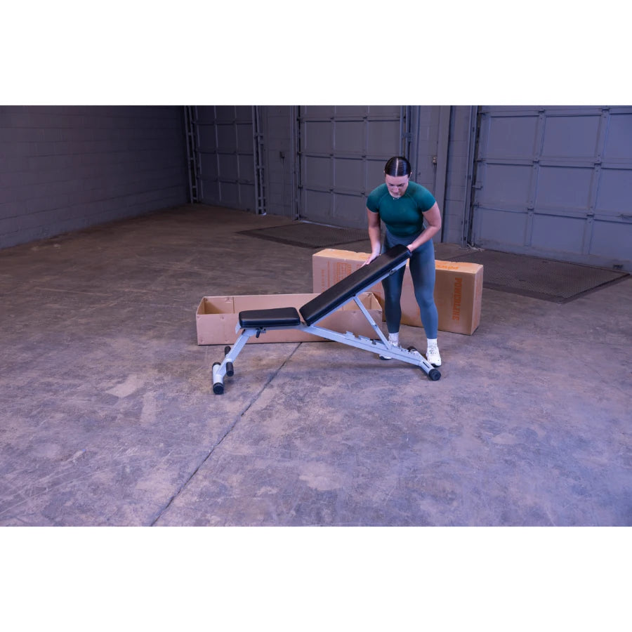 A person in athletic wear adjusting a weight bench in a spacious garage with cardboard boxes in the background.