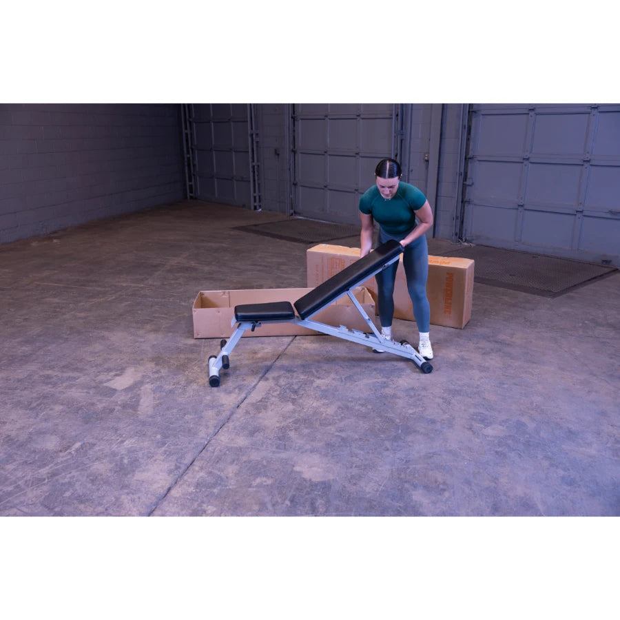 A woman in a green shirt and gray leggings adjusts an exercise bench in a spacious indoor area with cardboard boxes in the background.