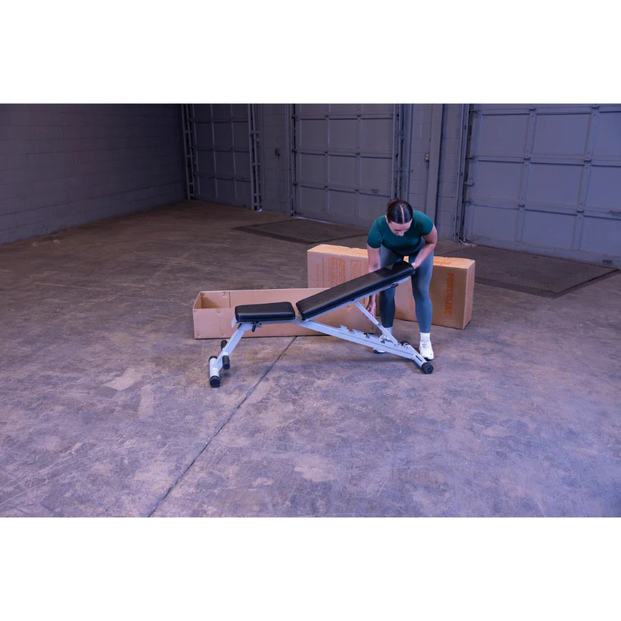 A person adjusting a workout bench in a spacious, unfinished room with cardboard boxes in the background.