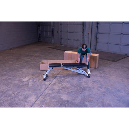 A person wearing a green shirt is assembling a workout bench inside a spacious garage, with two cardboard boxes in the background.