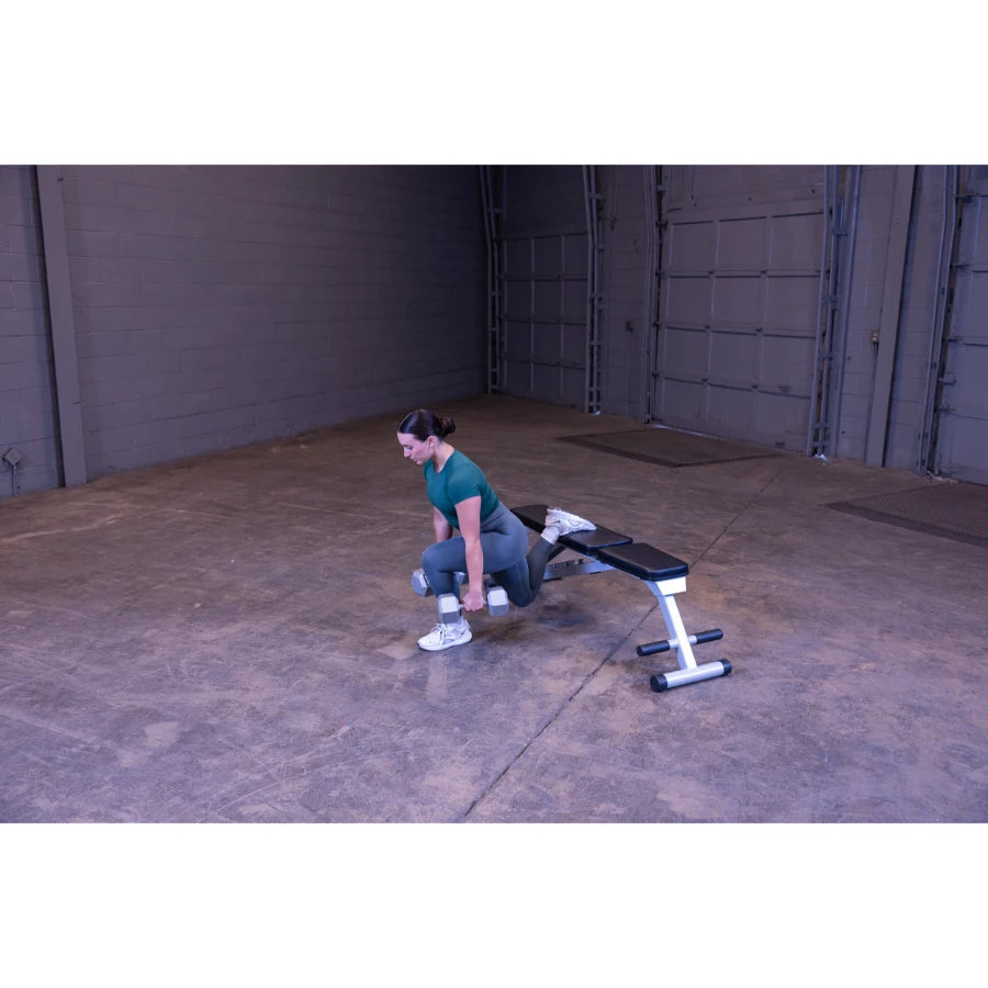 A woman in a green athletic outfit performs a dumbbell exercise in a spacious gym setting, using a bench for support.