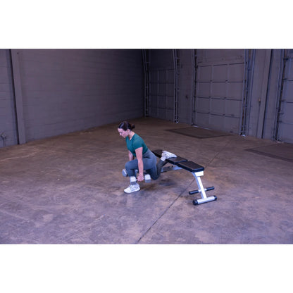 A woman in a green athletic outfit performs a dumbbell exercise in a spacious gym setting, using a bench for support.