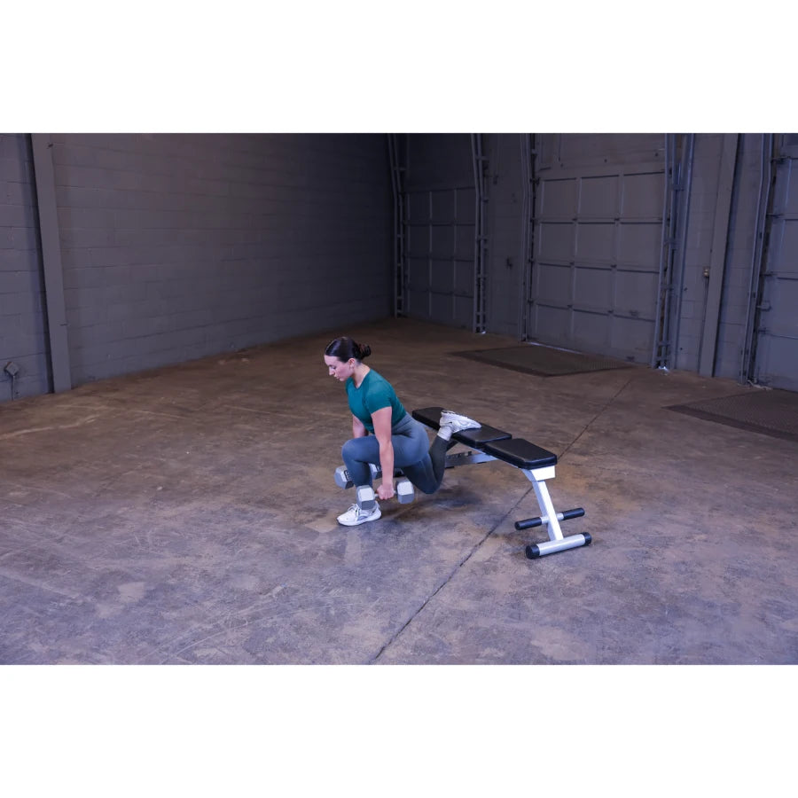 A woman in a green workout top and gray leggings performs a dumbbell exercise in an empty gym space, using a bench for support.
