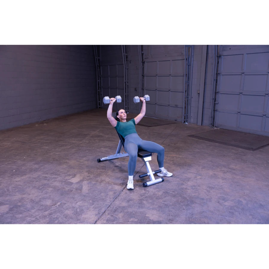 A person performing a dumbbell press while seated on a weight bench in a spacious gym or training area, wearing a green top and gray leggings.