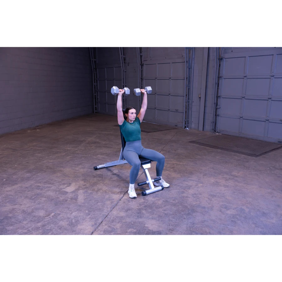 A person sitting on a weight bench in a gym, performing an overhead dumbbell press with both arms raised, wearing a green top and gray leggings.