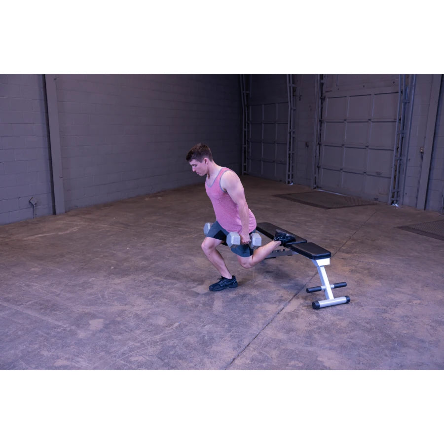 A man in a pink tank top and black shorts performs a lunge exercise with dumbbells in a gym setting, using a bench for support.