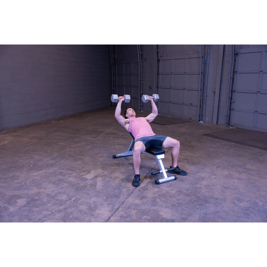 A person performing a dumbbell bench press exercise on a weight bench in an indoor gym setting. The individual is wearing a pink tank top and black shorts, lifting dumbbells above their chest.