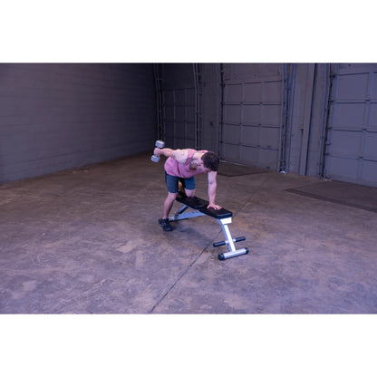 A man in a pink tank top and shorts performs a bent-over dumbbell row exercise on a weight bench in a spacious gym setting.