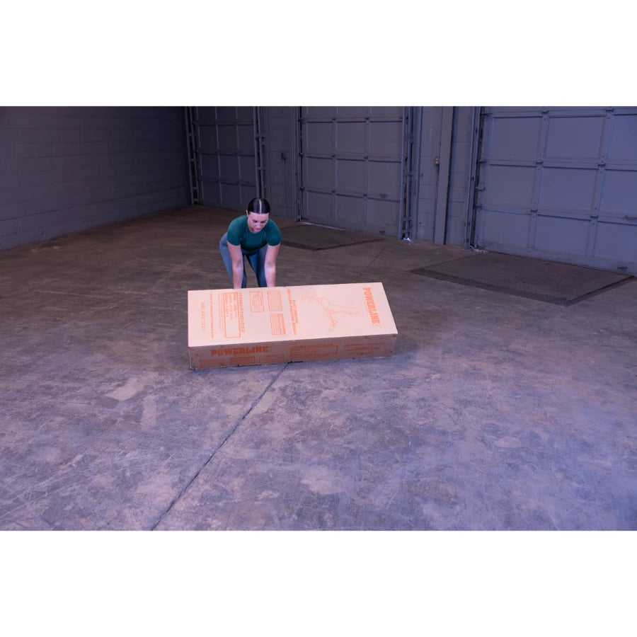 A person in a green shirt is bending down to lift a large cardboard box labeled POWERLINE in a spacious, empty warehouse setting.