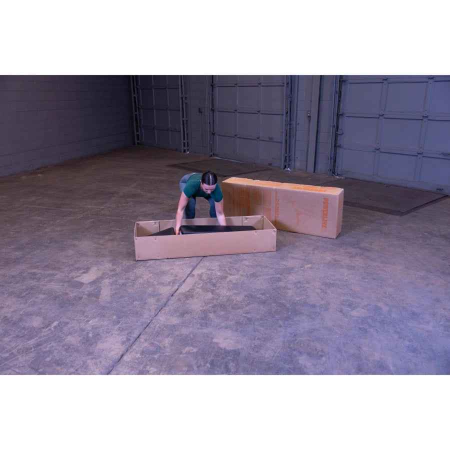 A person in a green shirt is leaning over a cardboard box, preparing to remove an item from it, with another cardboard box in the background, inside a spacious industrial setting.