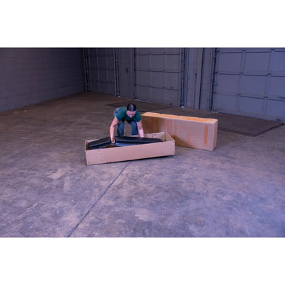 A person in a green shirt is leaning over a cardboard box, unpacking black items inside it, in a spacious, empty warehouse with concrete flooring. Another cardboard box is visible in the background.
