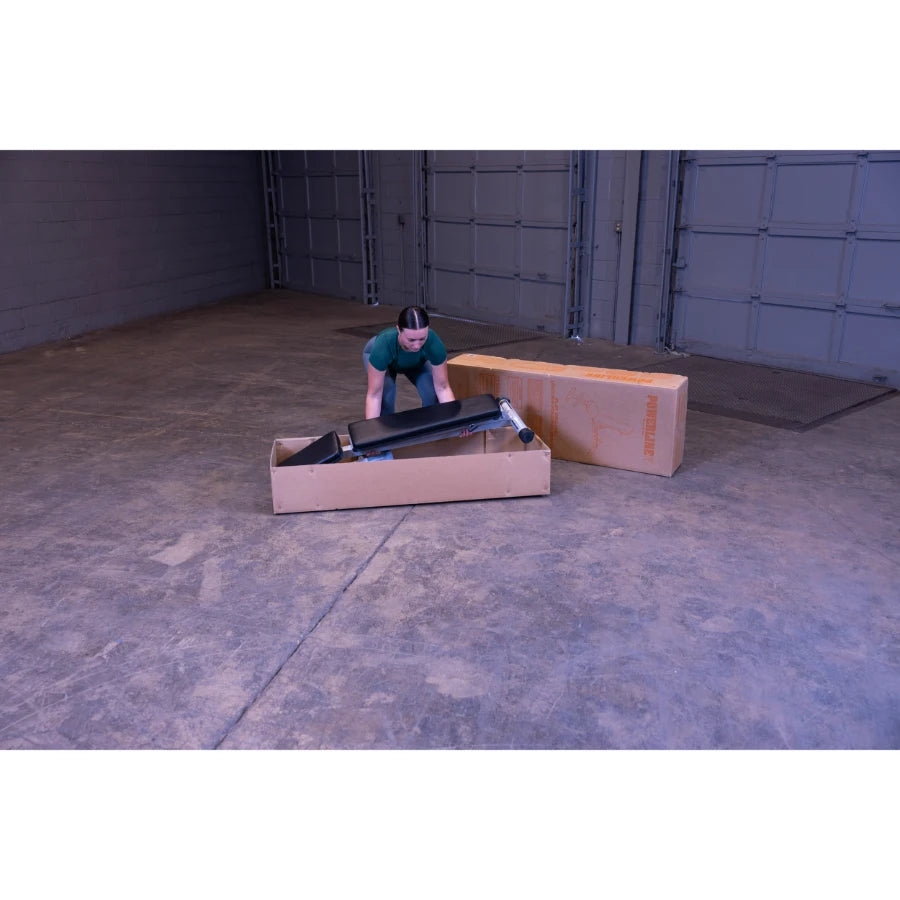 A person in a green shirt is unpacking a black item from a cardboard box in a spacious, empty warehouse with gray walls and a concrete floor.