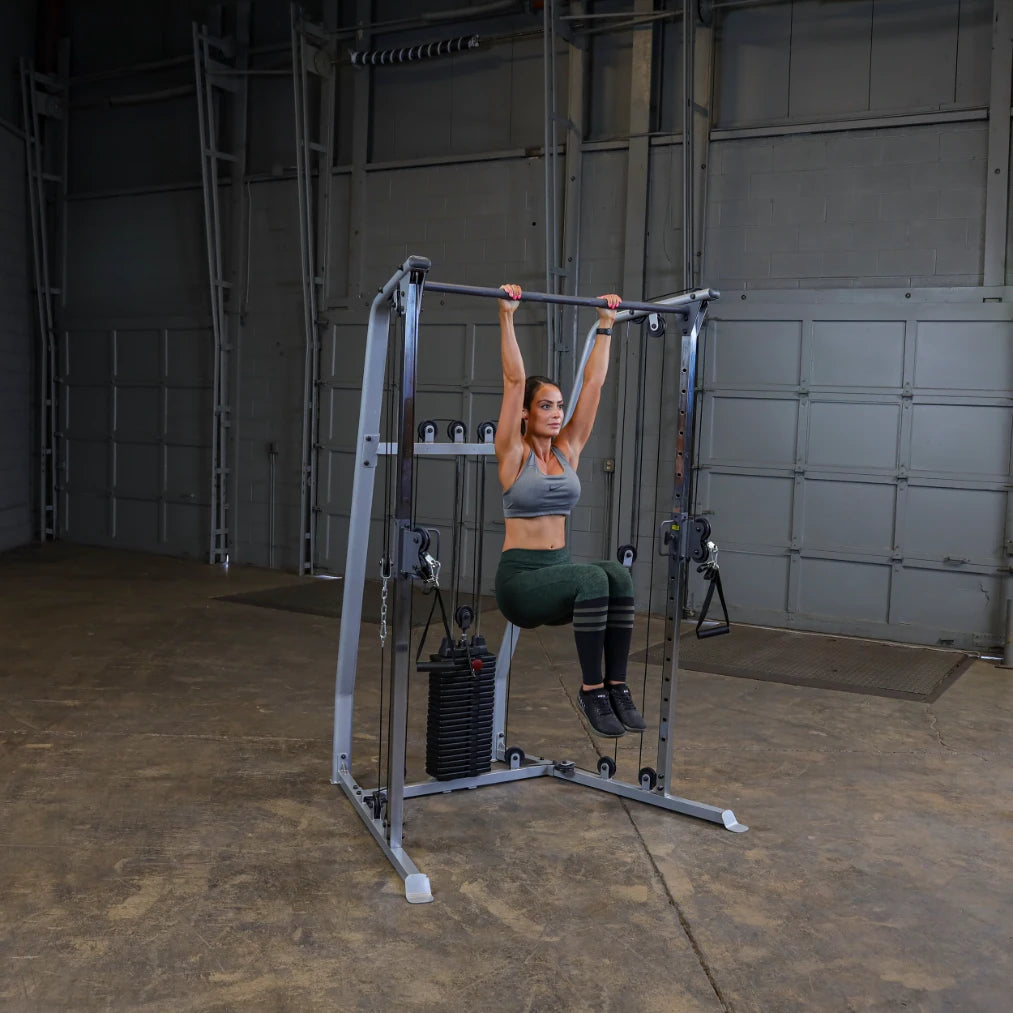 A woman performing an exercise on a multi-gym machine, hanging from a pull-up bar in a spacious indoor gym setting.