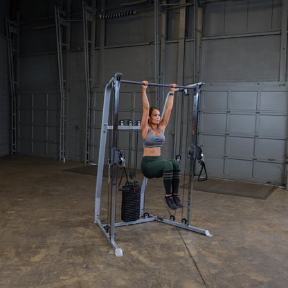 A woman performing an exercise on a multi-gym machine, hanging from a pull-up bar in a spacious indoor gym setting.