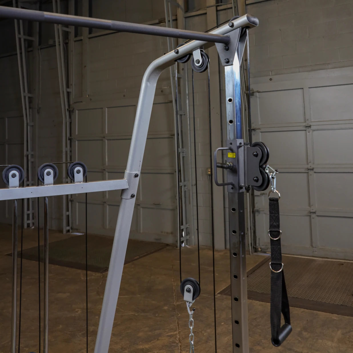 Close-up of a gym cable machine, featuring pulleys and a strap attachment, set against a backdrop of a spacious, industrial-style workout area.
