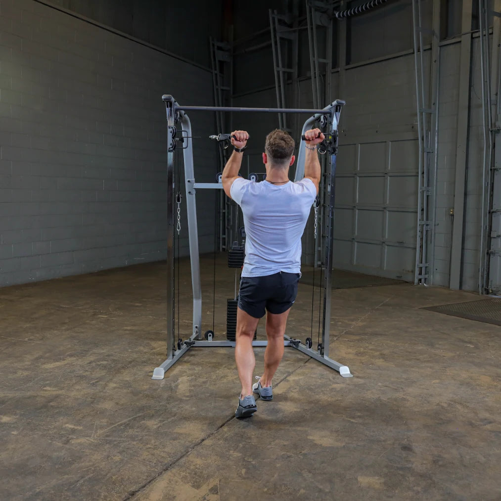 A person performing a cable exercise in a gym, facing away from the camera, with hands gripping the cable handles above shoulder height. The gym has a concrete floor and a minimalistic, industrial background.