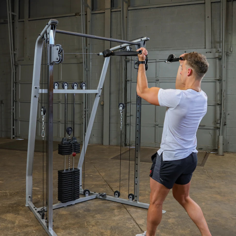 A person in a white t-shirt and black shorts is using a cable machine in a gym, performing a pull-down exercise. The gym has a spacious, industrial feel with gray walls.