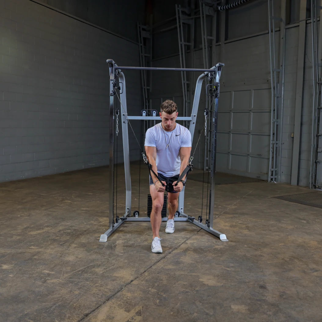 A person in a white athletic shirt and black shorts performs a cable exercise in a gym, using a cable machine with adjustable pulleys. The environment features a concrete floor and a plain wall in the background.