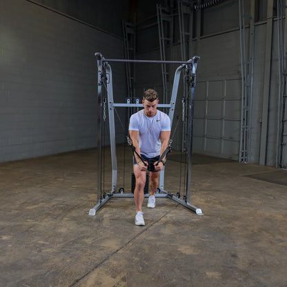 A person in a white athletic shirt and black shorts performs a cable exercise in a gym, using a cable machine with adjustable pulleys. The environment features a concrete floor and a plain wall in the background.