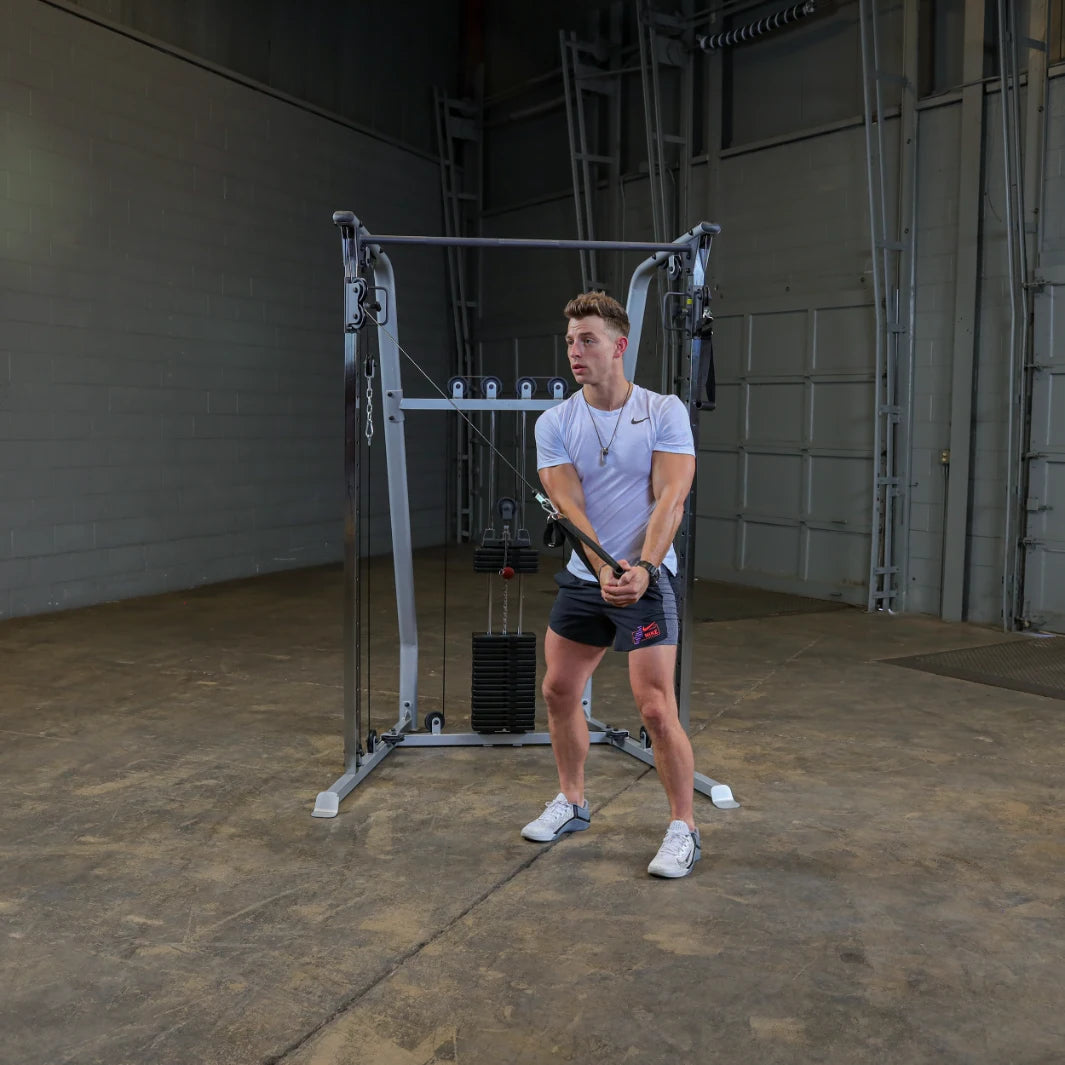 A young man in a white t-shirt and shorts is performing a cable exercise in a gym setting, using a cable machine with weights. He is focused and positioned to pull the cable towards him.