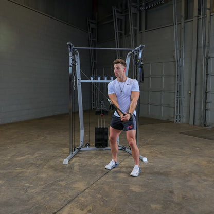 A young man in a white t-shirt and shorts is performing a cable exercise in a gym setting, using a cable machine with weights. He is focused and positioned to pull the cable towards him.