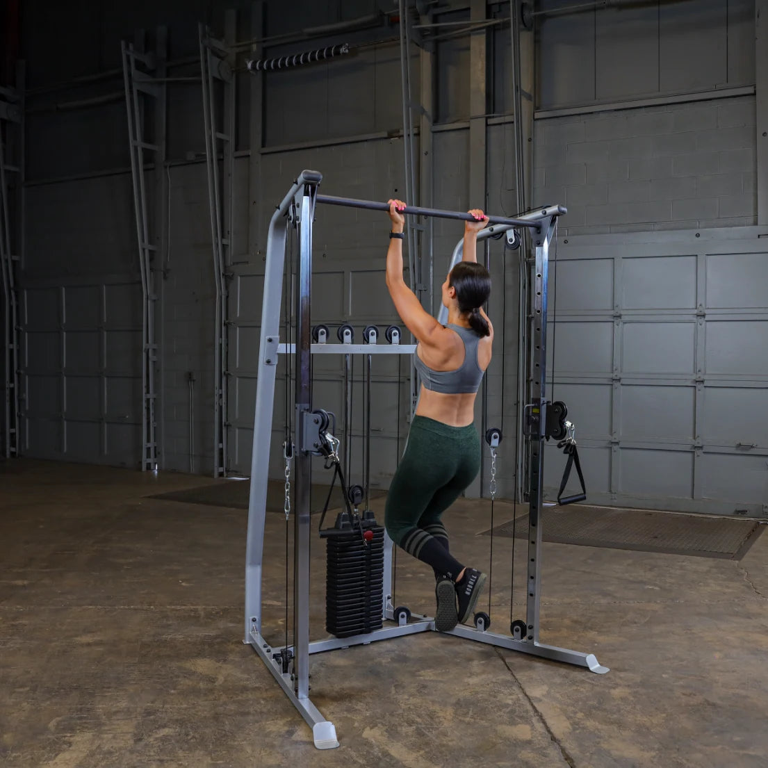 A woman performing a pull-up on a gym machine in a spacious indoor setting, wearing a sports bra and leggings, with weights visible at the base of the machine.