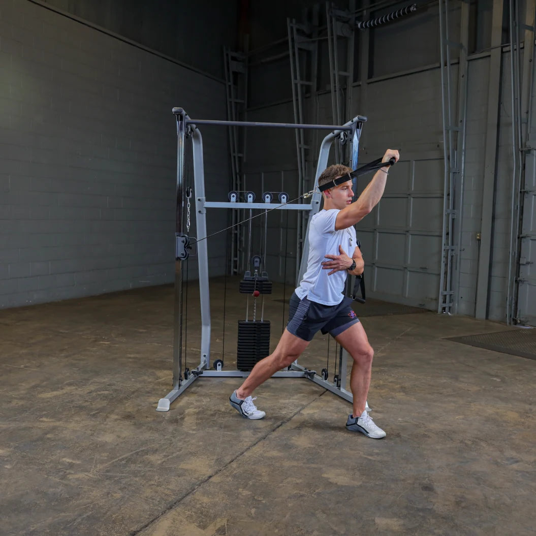 A man performs a cable exercise in a fitness gym, using a cable machine. He is wearing a headband and athletic clothing, demonstrating a dynamic movement with one arm extended forward while stabilizing himself with the other hand.