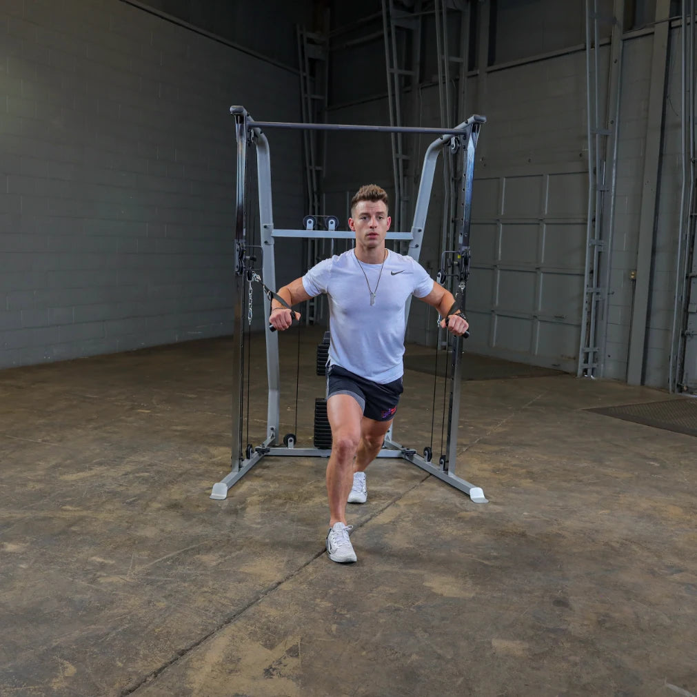 A young man wearing a white t-shirt and black shorts performs a workout using a cable machine in a spacious gym with concrete flooring.