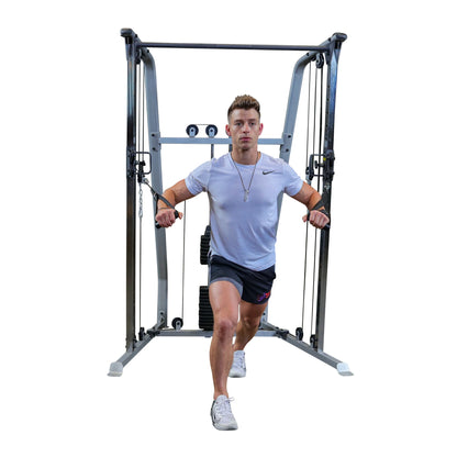 A man in a white t-shirt and black shorts is performing a cable exercise in a gym, using a cable machine with weights. He is in a lunge position, holding the cables with both hands, focused on his workout.