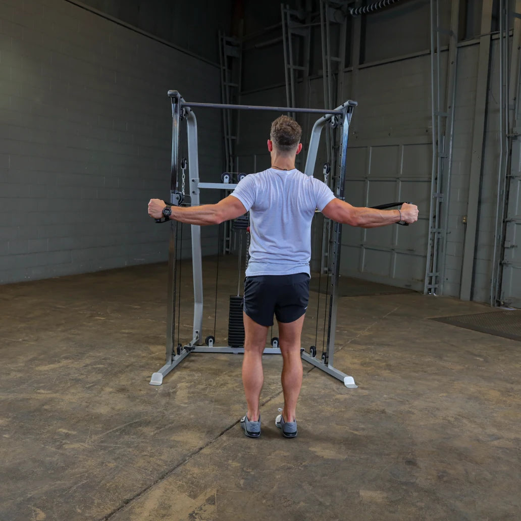 A person performing a cable exercise in a gym, standing with arms extended to the sides, wearing a gray t-shirt and black shorts, in a spacious workout area with a concrete floor.