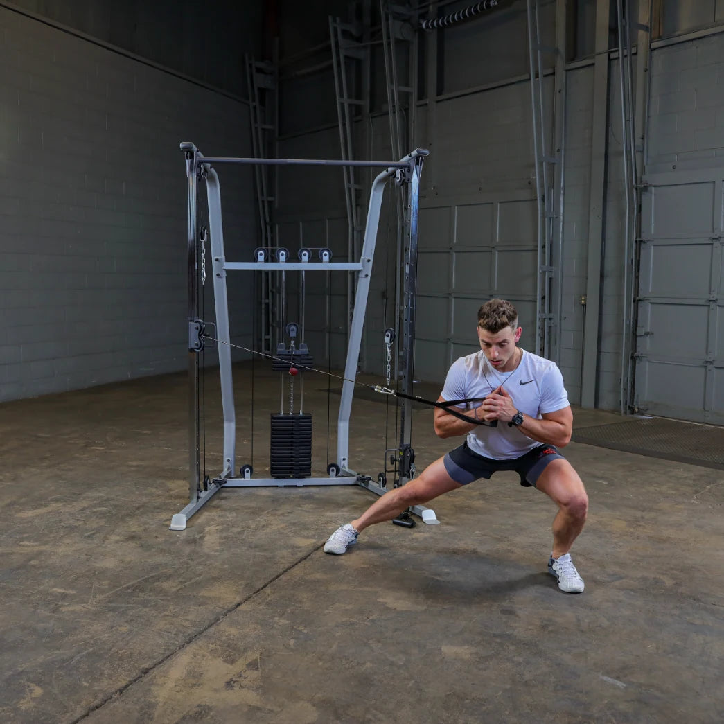 A young man in a gym performing a resistance band exercise, squatting to the side while holding a cable attachment, with a cable machine in the background.