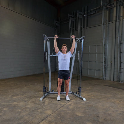 A young man wearing a white t-shirt and black shorts is performing an exercise on a cable machine, gripping the handles above his head in a gym setting with a concrete floor and gray walls.
