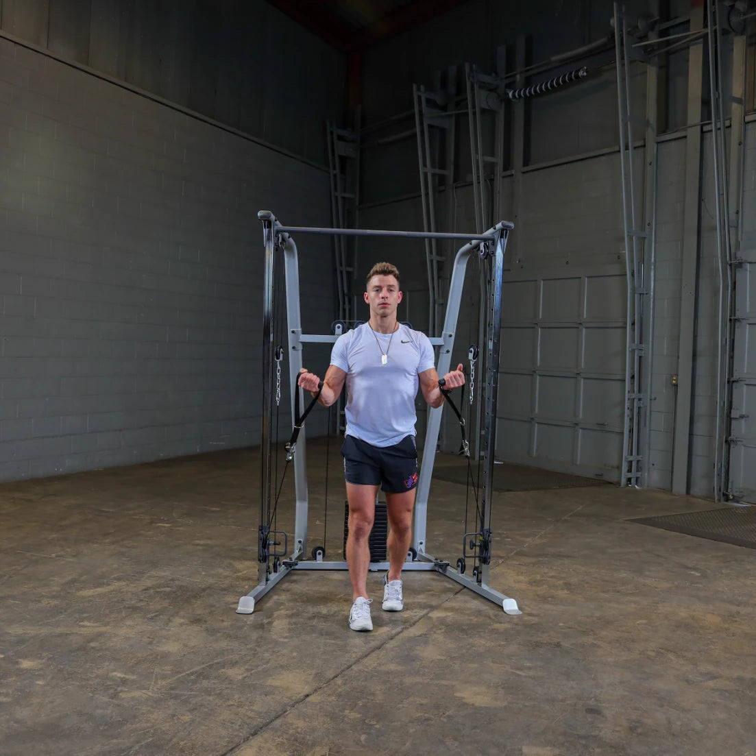 A person standing in a gym setting, using a cable machine for strength training. They are wearing a short-sleeved shirt and shorts, holding the handles of the machine with both hands. The background features gray walls and gym equipment.